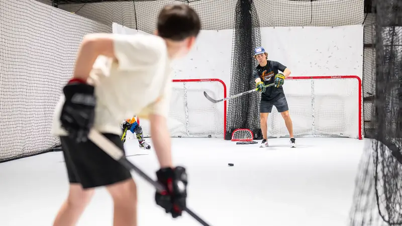 A young player loads up a shot on the synthetic ice lane while a coach tends the net